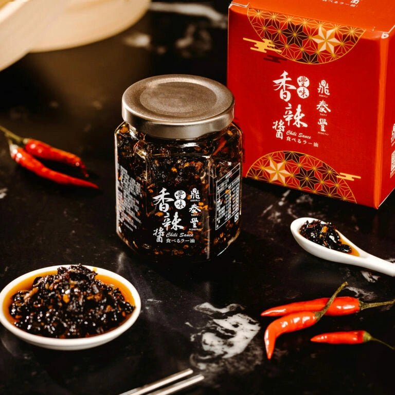 Jar of black bean sauce with a spoonful on a dark surface, next to red chili peppers and a red box.